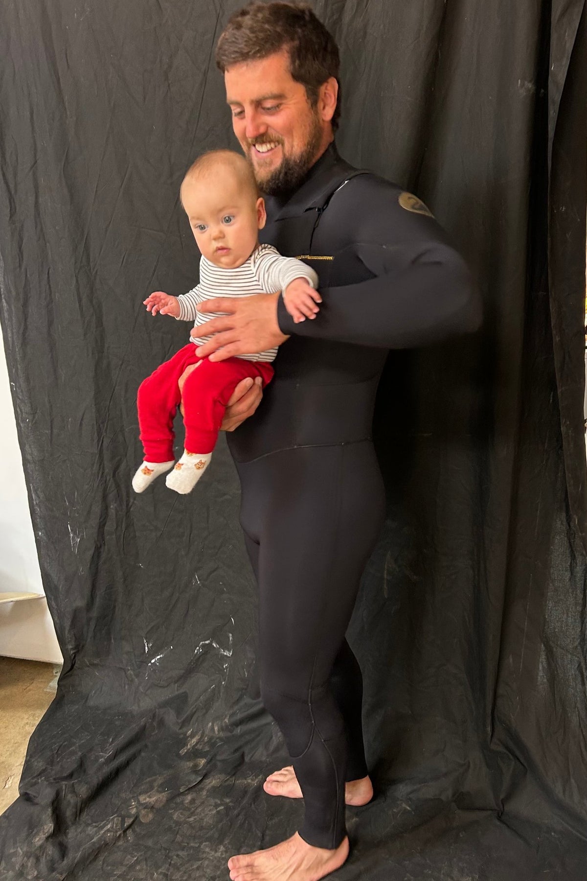 Black Duck Chest Zip 3/2mm Steamer full-length wetsuit worn by a surfer holding a baby - Moonshine Surfboards
